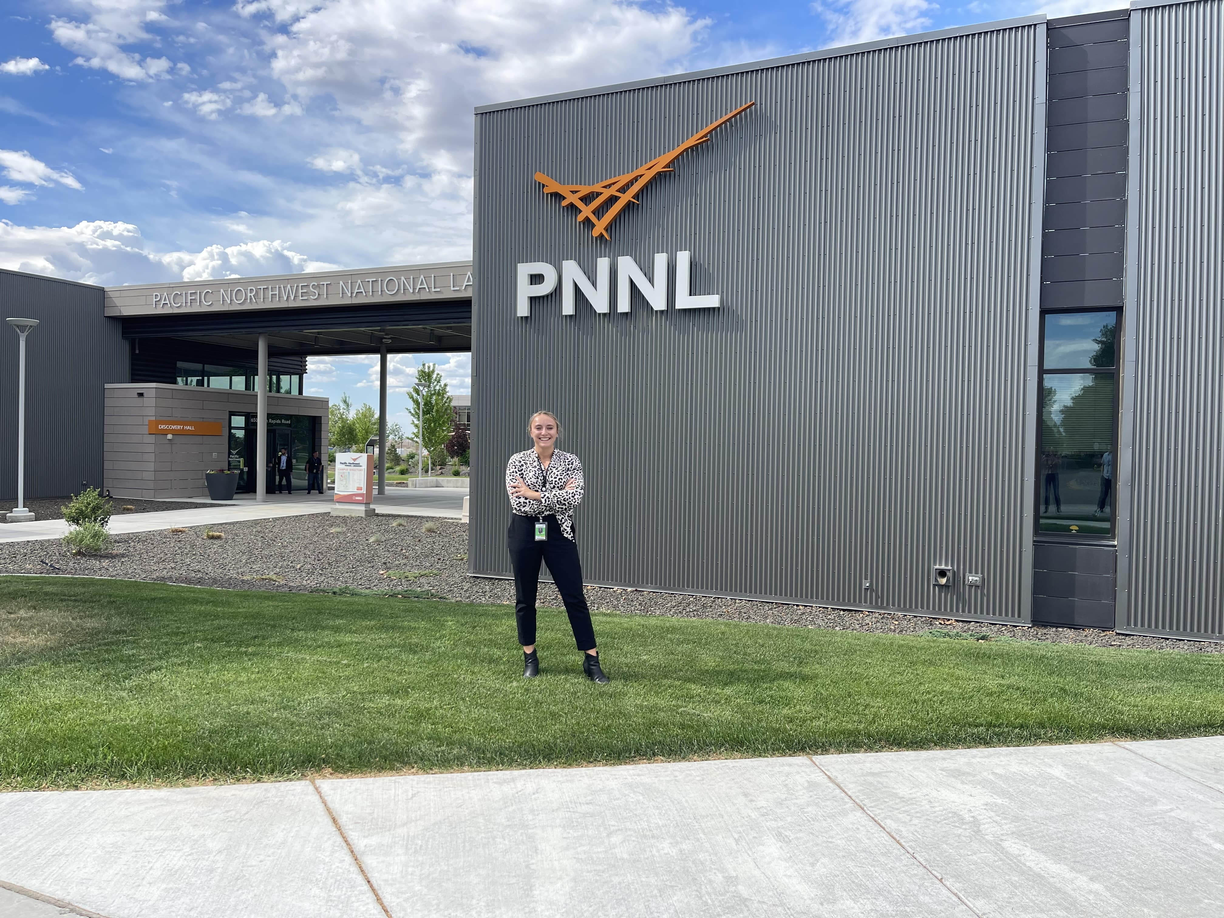 A person standing on grass in front of a gray building which has the letters PNNL on the outside. 