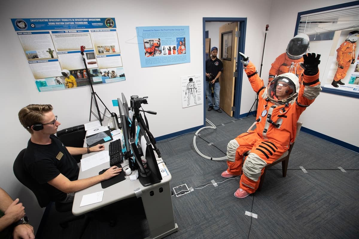A student wearing an orange space suit with raised arms in front of a student sitting at a computer.