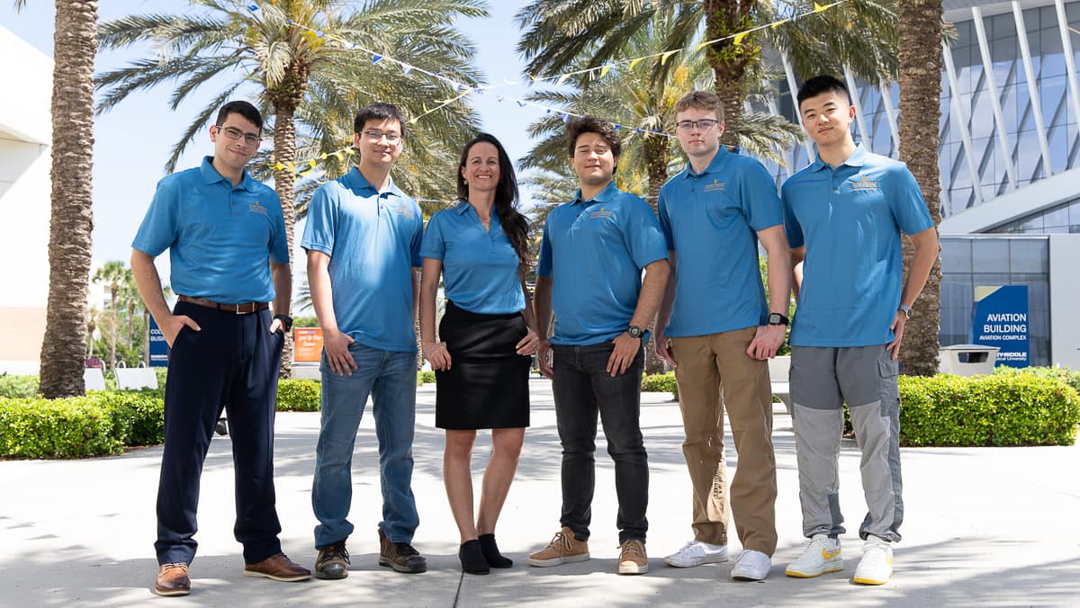 Five men and one woman wearing blue polos stand in a line in front of a glass building and palm trees