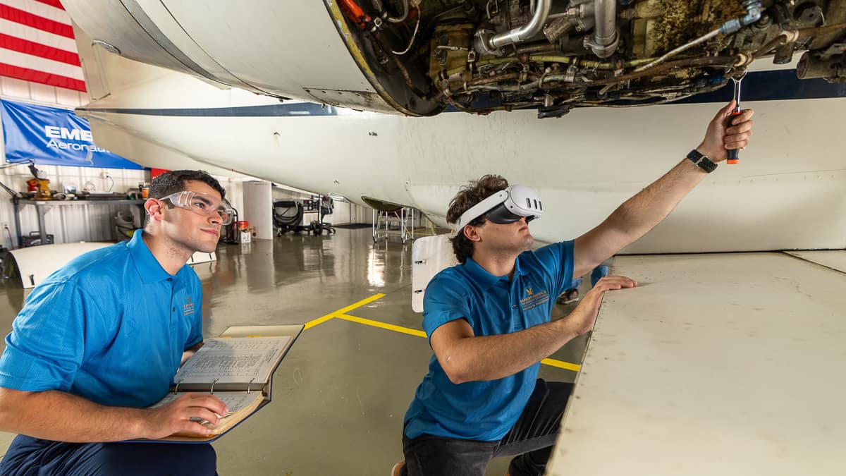 A man stands behind another man wearing a virtual reality headset and placing a wrench near the underside of a jet
