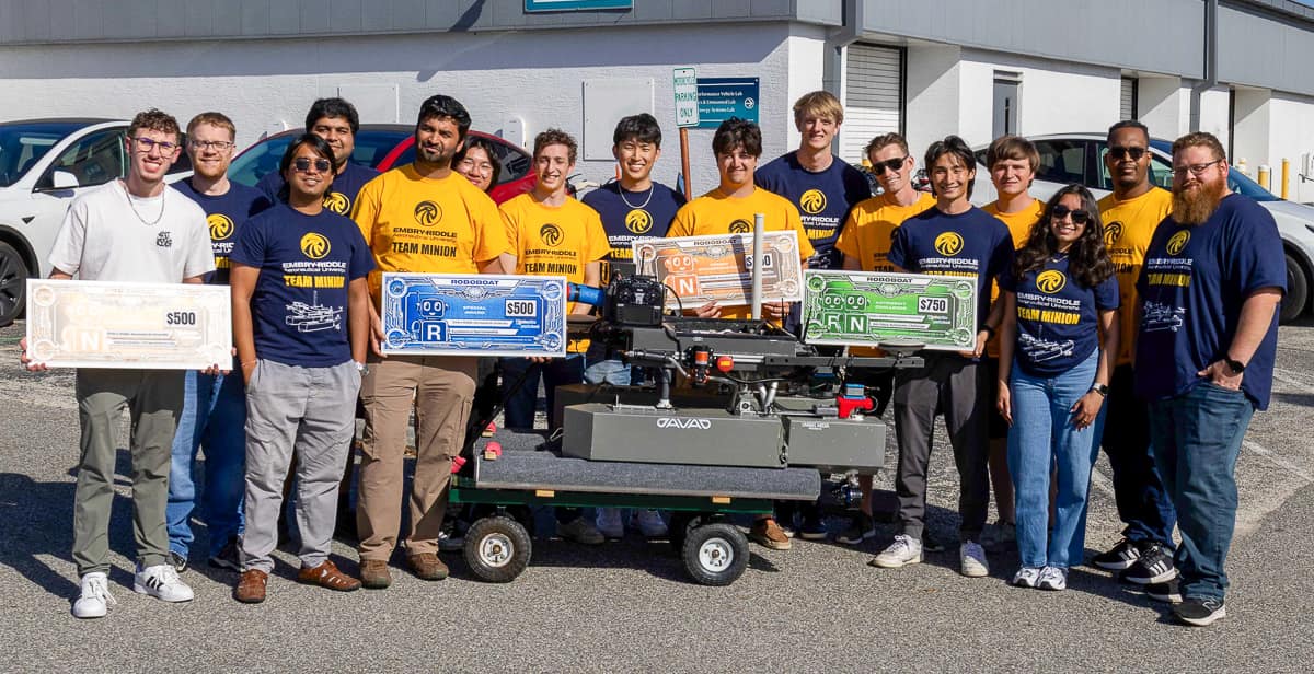 Fifteen people in blue and yellow shirts hold large checks in front of robotic boat
