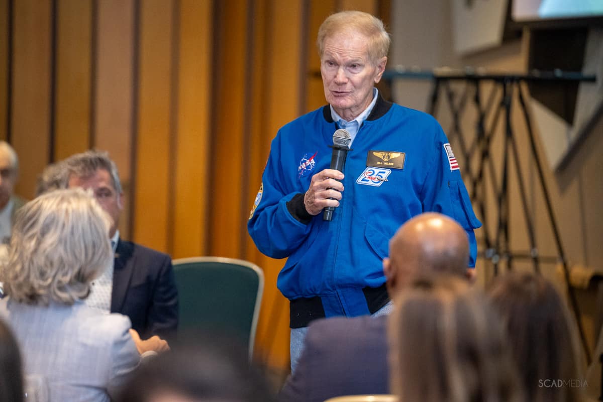 Man in NASA jacket holds microphone in crowded auditorium