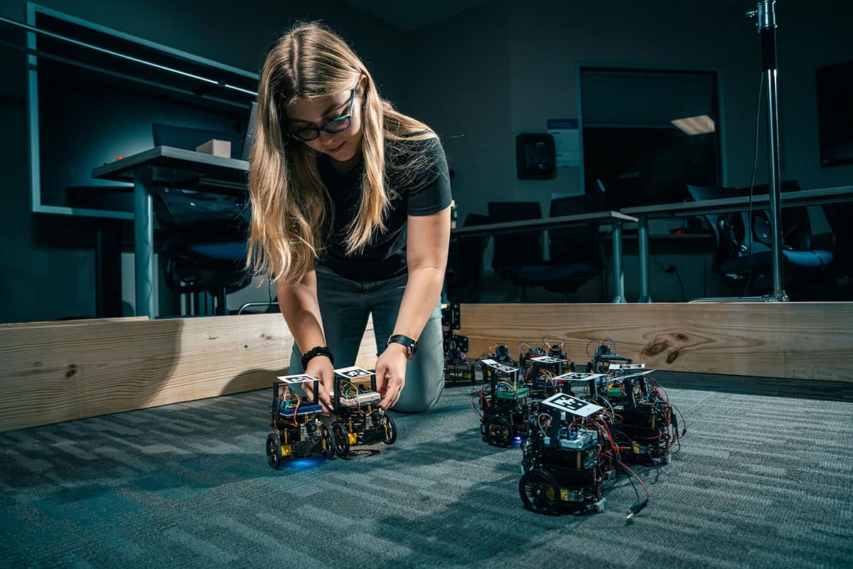 Student researcher Noa Teed, a senior majoring in Software Engineering, adjusts robotic vehicles programmed to interact at timed intervals and change each other's internal codes
